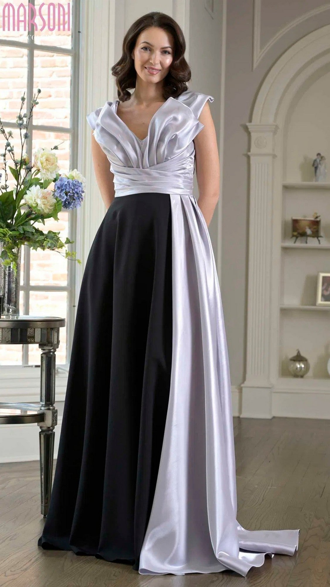 A woman smiles indoors wearing the Marsoni M345 Long Formal Dress in Black Silver, with flowers and shelves in the background.